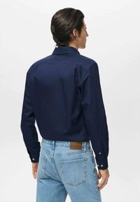 Camicia a maniche lunghe blu navy con colletto strutturato e polsini con bottoni, abbinata a jeans in denim azzurro chiaro dotati di una patch in pelle marrone.