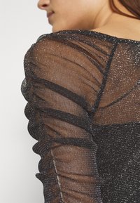 Svart genomskinlig topp med glitter, med rynkade ärmar och en figurnära kropp. Tyget har en slät textur och subtil lyster. Inga distinkta mönster.