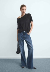 T-shirt negra de gran tamaño con un ligero diseño de un solo hombro, combinada con jeans de pierna ancha azules. La modelo sostiene un pequeño bolso negro y está de pie contra una pared clara.