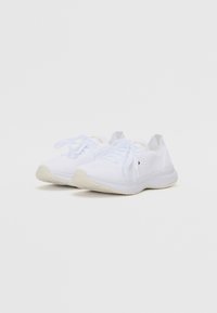White athletic shoes feature a mesh upper, flat white laces, and a textured off-white sole. Minimalistic design with no visible logos.