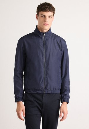Chaqueta bomber ligera de color azul marino con cremallera frontal, cuello alto y puños elásticos. Presenta un dobladillo fruncido y una textura suave.