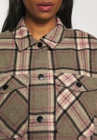 Chaqueta de camisa a cuadros en verde oliva con patrones en rosa y negro, que presenta un cuello con solapa, botones a presión negros y bolsillos frontales. Material de lana.