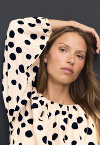 Krēmkrāsas blūze ar lieliem melniem punktiņiem un pūkainām piedurknēm. Izceltas pūstās kakla malas. Gluda, viegla auduma.