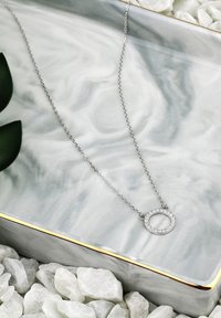 Sølv halskæde med en rund vedhæng prydet med små klare sten, som hviler på en marmor grå bakke med en gylden kant.