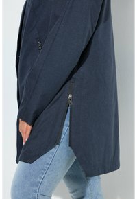 Cappotto blu navy con dettaglio zip laterale indossato sopra jeans azzurri, con mano appoggiata vicino all'anca.