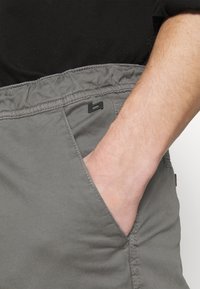 Sive kratke hlače s elastičnim pojasom, koje imaju bočni džep i mali crni detalj s logom. Materijal je gladak s mat završnom obradom.