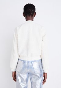 White textured bomber jacket with a cropped cut, ribbed cuffs, paired with shiny silver-blue metallic pants. View from the back.
