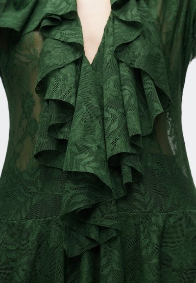 Πράσινο δαντελένιο φόρεμα με στρωμάτωση βολάν στο λαιμό και το μπούστο. Υφή με λουλουδάτο μοτίβο σε διάφανο ύφασμα. Βαθύ V ντεκολτέ.