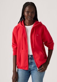 Hoodie rouge à zip avec deux poches avant, cordons de serrage et poignets côtelés, associé à un haut blanc et un jean bleu. Texture lisse, coupe décontractée.