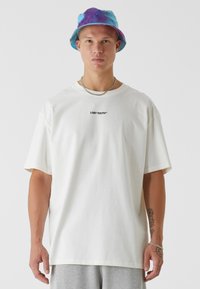 Biely bavlnený tričko s krátkymi rukávmi a čiernym nápisom "LOST YOUTH". Má voľný strih a hladkú textúru. Nosí sa s viacfarebným klobúkom.