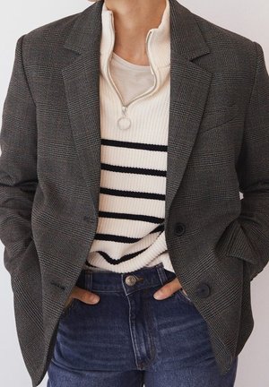 Person iført en grå ternet blazer over en hvid ribbet sweater med sorte striber og højtaljede blå jeans, hænderne i lommerne.