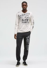 Beige Hemd mit langen Ärmeln und schwarzen Grafikdrucken, kombiniert mit dunkelgrauen Jogginghosen mit weißem Text, abgerundet mit schwarzen Sneakers.