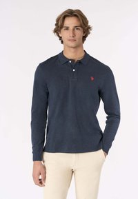 Polo shirt a maniche lunghe blu navy in misto cotone, con colletto e due bottoni. Logo rosso sul lato sinistro del petto. Indossato con pantaloni chiari.