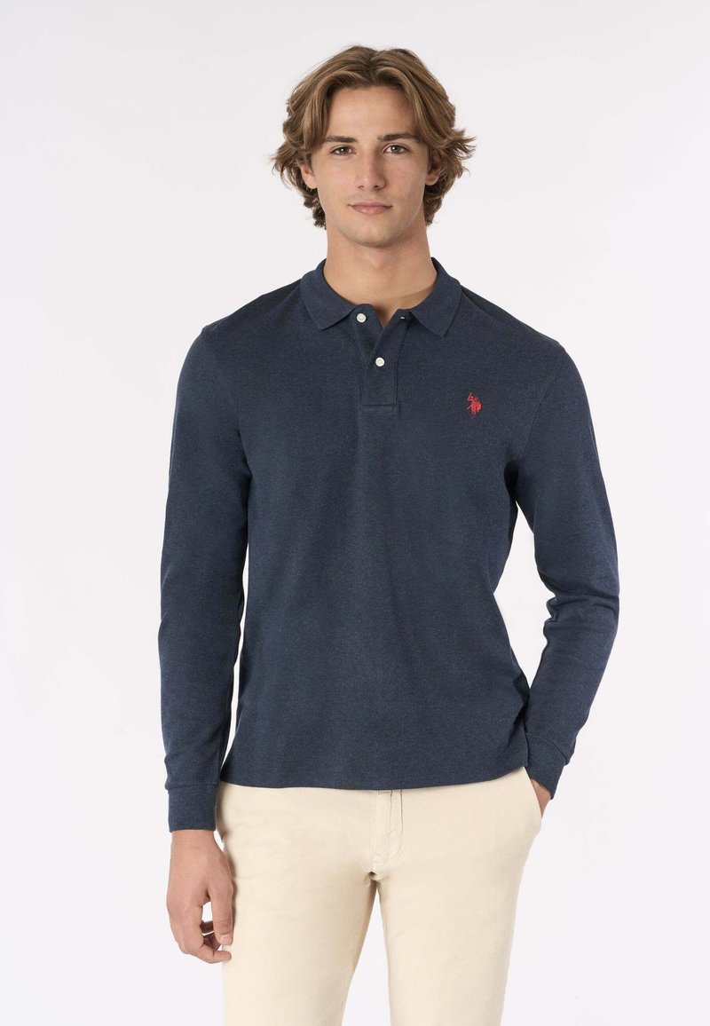 Polo shirt a maniche lunghe blu navy in misto cotone, con colletto e due bottoni. Logo rosso sul lato sinistro del petto. Indossato con pantaloni chiari.