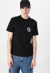 Black cotton t-shirt with a crew neck and a small white logo on the left chest, paired with blue denim cargo shorts.