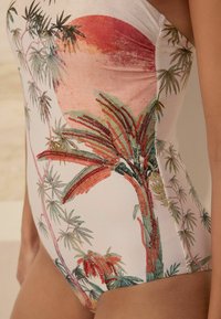 Enbitar swimsuit med tropiskt mönster av palmblad och röda accenter, tillverkad av ett mjukt, elastiskt tyg, i mjuka pastellfärger.
