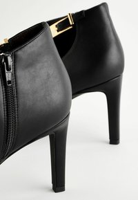 Stivali alla caviglia in pelle nera con texture liscia, chiusura con zip e fibbia con accenti dorati. Presentano un tacco alto e slanciato.