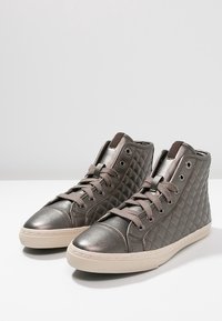 Metalické šedé vysoké tenisky mají prošívanou texturu, kulatou špičku a béžovou gumovou podrážku s plochými tkaničkami a kovovými očky.