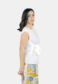 Top bianco senza maniche con un nodo in vita, abbinato a pantaloni con fantasia che presenta foglie blu e gialle e forme geometriche.