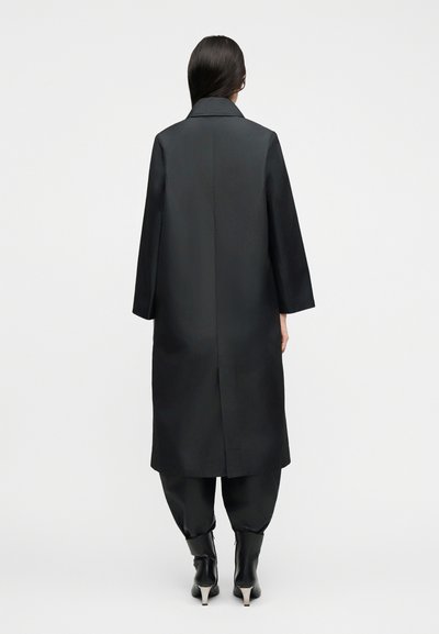 Γυναίκα με μακριά σκούρα μαλλιά φορά μαύρο μακρύ παλτό, φαρδιά μαύρη παντελόνα και μαύρα μποτάκια με τακούνι, στέκεται γυρισμένη με την πλάτη σε λευκό φόντο.