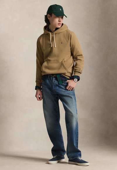 Mladý muž oblečený v olivovo zelenom pulóvri s kapucňou, zelenej šiltovke, modrých džínsoch a teniskách stojaci pred neutrálnym pozadím s rukami v vreckách.