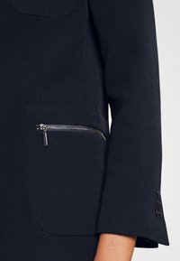 Veste noire sur mesure avec une poche zippée, un design structuré, un tissu texturé et des boutons en accent sur la manche.