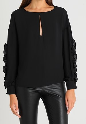 Blouse noire à manches longues avec encolure en trou de serrure à l'avant et manches transparentes à volants, associée à un pantalon en cuir noir porté par une femme.