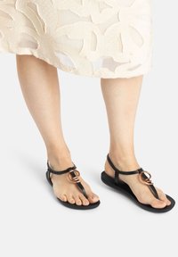 Black thong sandals feature a flat sole, minimalist straps, and a circular copper accent. The design is sleek and functional.