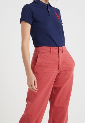 Person trägt ein dunkelblaues Polohemd mit rotem Logo und hoch taillierte, locker sitzende rote Hose, stehend vor einem einfarbigen Hintergrund.