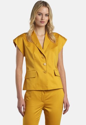 Blonde vrouw draagt een mosterdgele mouwloze blazer met drie knopen en bijpassende op maat gemaakte broek tegen een witte achtergrond.