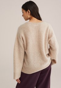 Gai bežs oversize džemperis, izgatavots no mīksta materiāla, ar noapaļotu kakla izgriezumu un ribotām malām, apvienots ar tumši violetiem biksēm.