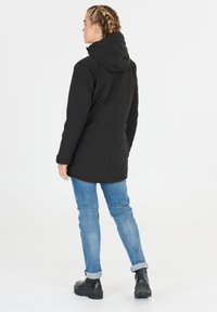 Sort hættejakke med skræddersyet pasform, der har en glat tekstur, forlænget design og sidelommer, parret med lyseblå jeans og sorte støvler.