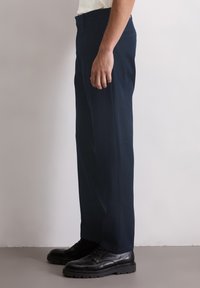 Pantalon bleu marine en tissu texturé, coupe droite, avec une poche latérale discrète, porté avec des chaussures noires à lacets.