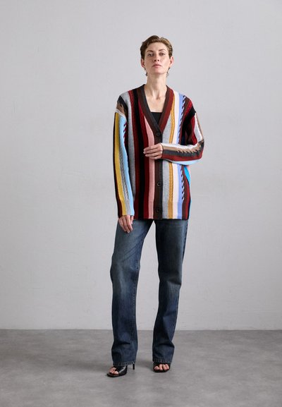 Strickjacke mit mehrfarbigen vertikalen Streifen in Rot, Blau, Gelb und Braun; mit V-Ausschnitt und Knöpfen. Kombiniert mit geraden Jeans.
