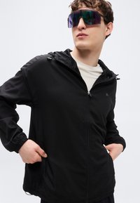 Μαύρο μπουφάν με φερμουάρ, με κουκούλα από δίχτυ, ελαστικά μανίκια και τσέπες. Το μοντέλο φοράει μεγάλα σκουρόχρωμα γυαλιά ηλίου. Λείος υφασμάτινος ιστός και εφαρμογή.