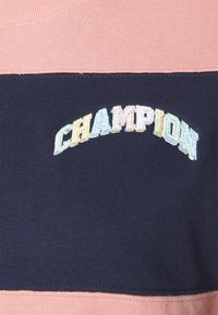 Wielokolorowy haftowany napis "CHAMPION" na granatowym tle, otoczony jasnoróżowym materiałem powyżej i poniżej.