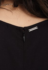 Vêtement noir avec une fermeture éclair à l'arrière, doté d'une petite étiquette en argent avec le logo "VOLCOM" fixé près de l'encolure.