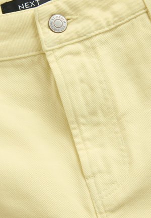 Close-up van gele denimstof met knoop- en stikdetails, en een zichtbaar label met de tekst "NEXT DENIM."