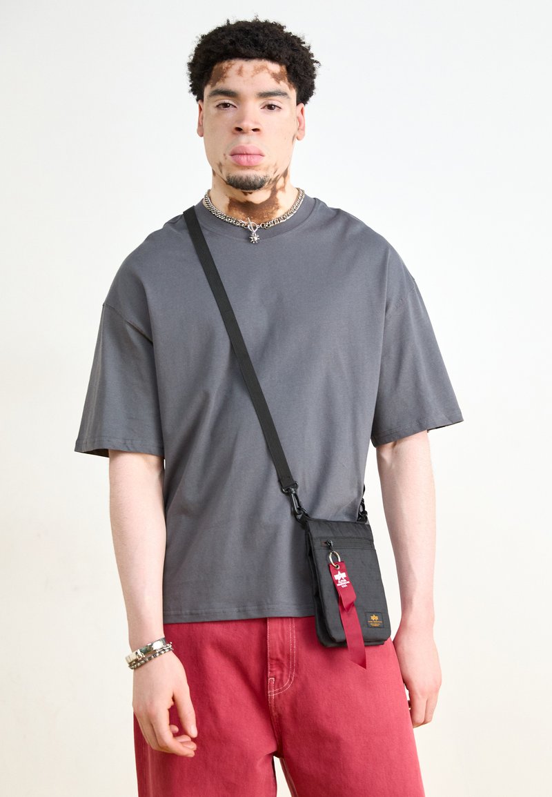 T-shirt cinza de manga curta combinado com calças largas vermelhas. Saco crossbody preto com uma etiqueta vermelha e detalhes em metal. Colar de corrente prateada.