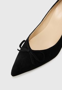 Ballerines en daim noir avec un bout pointu et un petit nœud en accent à l'avant. Doublure intérieure lisse, talon minimal et design classique.