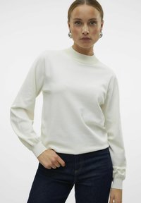 Sweatshirt blanc à manches longues avec un col haut et des poignets côtelés. Texture douce, design minimaliste, associé à un jean bleu marine.