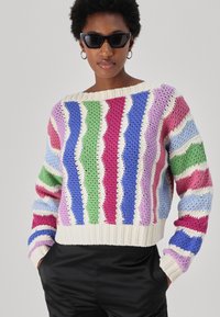 Häkelpullover mit weitem Ausschnitt, mit wellenförmigen Streifen in Pink, Lila, Blau, Grün und Creme, kombiniert mit schwarzen Hosen.