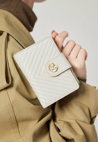 Portefeuille matelassé blanc avec un fermoir circulaire doré et des détails zippés, tenu dans une main. L'arrière-plan présente un tissu beige.