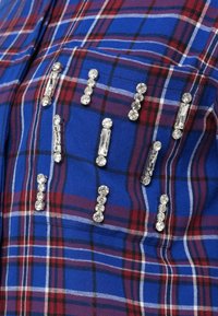 Kreklu ar apakšmalu, kuram ir zils, sarkans un balts kliņģerīšu raksts. Rotāts ar rhinestones vertikālos rakstos, pievienojot spīdumu un tekstūru.