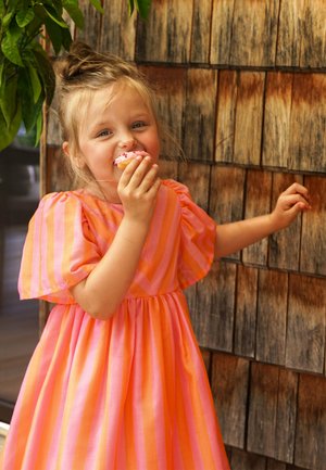 Rosa-orange gestreiftes Kleid mit Puffärmeln und geraffter Taille. Das Kind hält einen Cupcake mit rosa Zuckerguss und Streuseln.