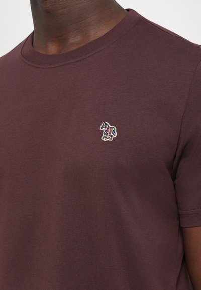 Man in een donkerpaarse T-shirt met een klein geborduurd meerkleurig zebra logo op de linkerborst.