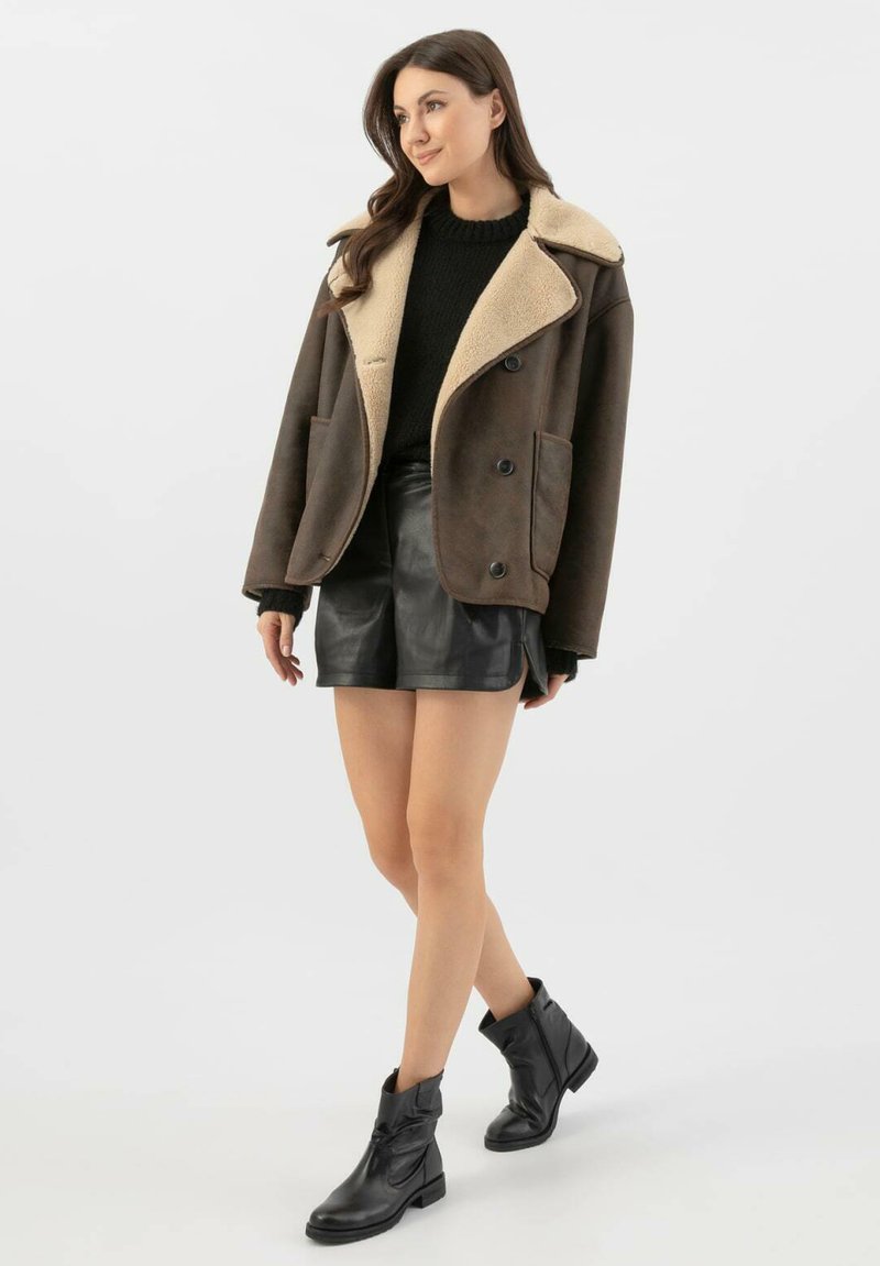 Hnědý ovčí kožich s krémovým límcem, černý svetr a černé kožené kraťasy. Modelka má na sobě černé kotníkové boty a stojí na neutrálním pozadí.