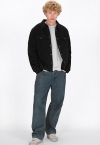 Giacca nera con bottoni e due tasche sul petto sopra una maglietta grigia, abbinata a jeans blu larghi e sneakers grigie chiare. Look casual.