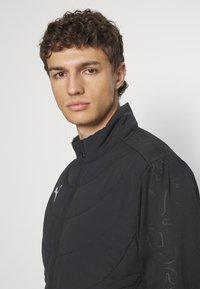 Schwarze Zip-Jacke mit hohem Kragen, strukturierten Schulterakzenten und einem dezenten Muster am Ärmel. Puma-Logo in Grau auf der Brust.