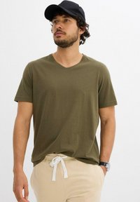 T-shirt in cotone verde oliva con scollo a V, maniche corte e vestibilità rilassata, abbinata a pantaloni beige con coulisse e un berretto nero.